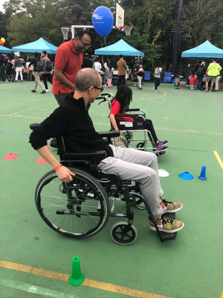 A girl and a man are competing in a wheelchair race, behind the girl her father can be seen giving her encouraging her