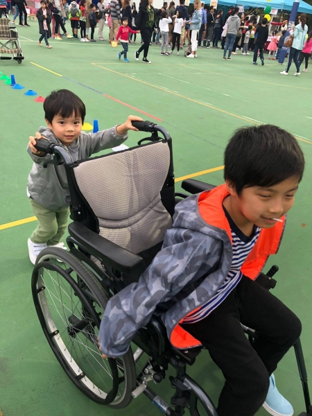A boy can be seen seated in a wheelchair while his little brother is behind him helping him push the wheelchair.