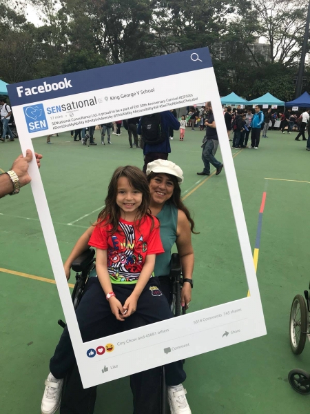 A mum sitting in a wheelchair can be seen posing behind the SENsational Facebook post photo frame with her daughter on her lap