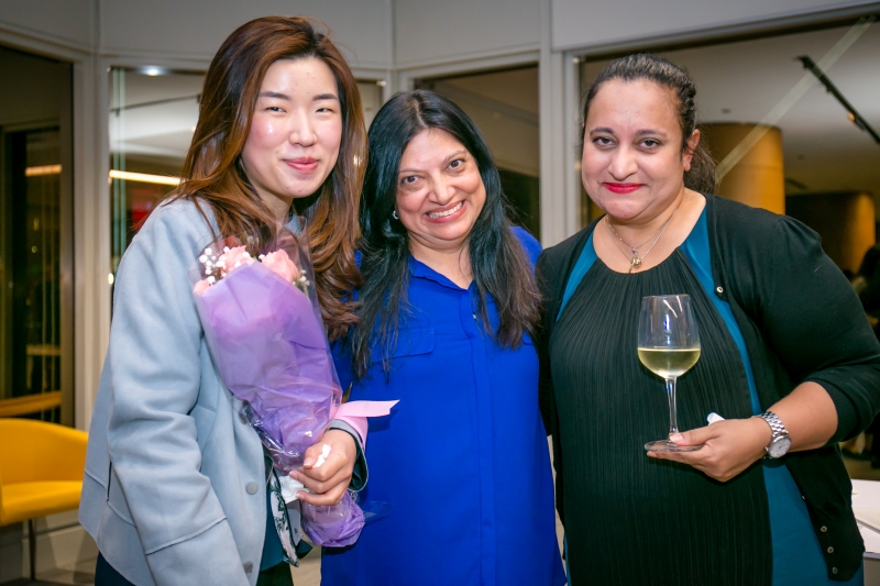 3 women in business attire are grouped together for a photo. 1 of the woman can be seen holding a bouquet whilst another has a glass of wine.